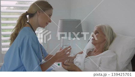 Leaning nurse wearing blue scrubs offering glass of water to patient on hospital bed in room Leaning nurse wearing blue scrubs offering glass of water to patient on hospital bed in room 139425701