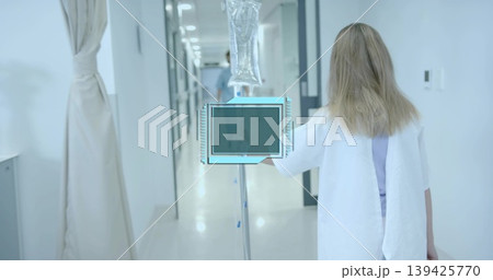 Walking patient with wristband in gown pushing IV drip stand down white corridor, sterile ward 139425770