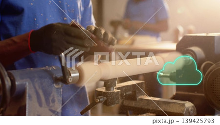 Craftsman wearing blue shirt and red wristband turning spindle on lathe in workshop, with shavings 139425793