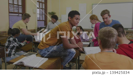 Leading student wearing mustard t-shirt sitting on wooden desk in classroom, with open textbooks Leading student wearing mustard t-shirt sitting on wooden desk in classroom, with open textbooks 139425834