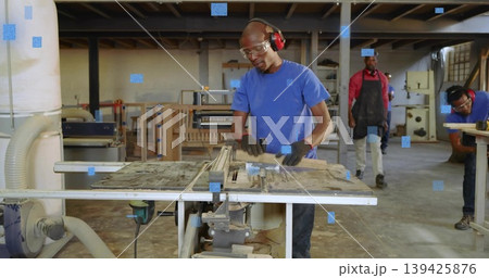 Operating carpenter feeding wooden board through table saw in workshop, with sawdust and gloves 139425876