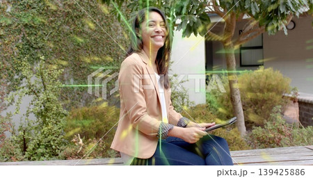 Holding tablet, Asian woman wearing beige blazer and jeans sitting on bench in landscaped courtyard Holding tablet, Asian woman wearing beige blazer and jeans sitting on bench in landscaped courtyard 139425886