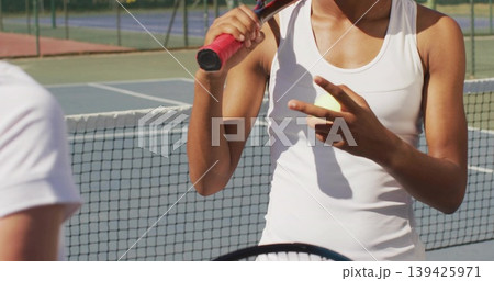 Signaling female tennis player holding racket near net on outdoor court, with fence and court lines 139425971
