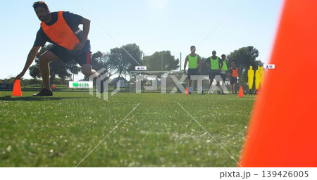 Lunging male athlete in orange bib reaching cone on turf, with goal, mannequins, fence, houses Lunging male athlete in orange bib reaching cone on turf, with goal, mannequins, fence, houses 139426005