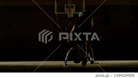 Competing adult basketball players jostling under gym hoop, spotlight casting shadows on jerseys Competing adult basketball players jostling under gym hoop, spotlight casting shadows on jerseys 139426008