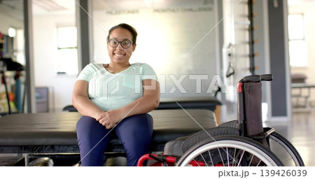 Sitting patient wearing mint T-shirt navy leggings on therapy table in clinic, near red wheelchair 139426039