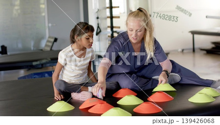 Reaching clinician in navy scrubs placing disc cones on large black padded mat while child watching Reaching clinician in navy scrubs placing disc cones on large black padded mat while child watching 139426068