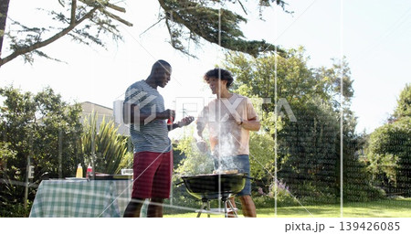 Grilling two men tending charcoal grill in yard holding sauce bottle and tongs near checked table Grilling two men tending charcoal grill in yard holding sauce bottle and tongs near checked table 139426085