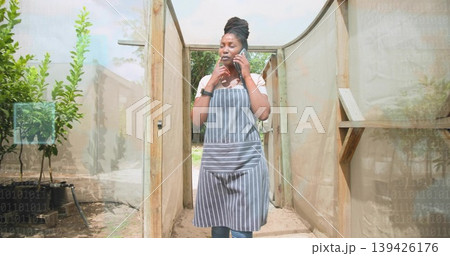 Holding smartphone, apron-clad gardener checking messages in mesh tunnel, with potted trees Holding smartphone, apron-clad gardener checking messages in mesh tunnel, with potted trees 139426176