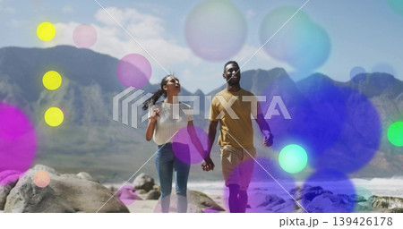 Walking woman and African American man holding hands at beach, with boulders, waves, mountain peaks Walking woman and African American man holding hands at beach, with boulders, waves, mountain peaks 139426178