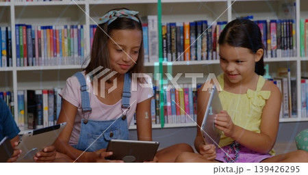 Sitting two girls in denim overalls, yellow bow top reading tablets in library, with bookshelves Sitting two girls in denim overalls, yellow bow top reading tablets in library, with bookshelves 139426295