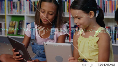 Reading two schoolgirls crosslegged using tablets on library carpet, with bookshelves backdrop 139426298