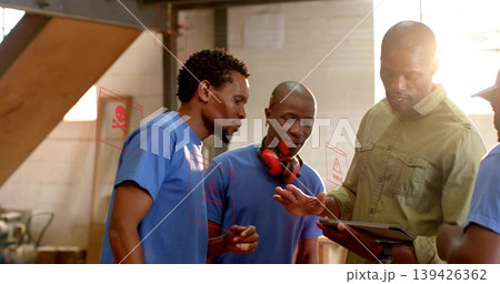 Examining tablet man in olive work shirt guiding blue tshirt workers in workshop with earmuffs Examining tablet man in olive work shirt guiding blue tshirt workers in workshop with earmuffs 139426362
