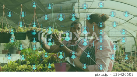 Plant caretakers wearing aprons examining hanging seedlings in greenhouse, with digital icons Plant caretakers wearing aprons examining hanging seedlings in greenhouse, with digital icons 139426505