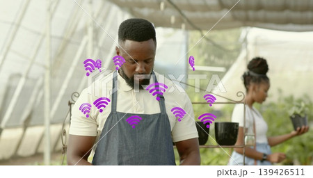 Horticultural worker wearing denim apron inspecting plant in greenhouse, with purple wireless icons 139426511