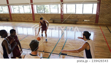 Dribbling adult male in maroon uniform moving across school gym court, holding orange basketball Dribbling adult male in maroon uniform moving across school gym court, holding orange basketball 139426543