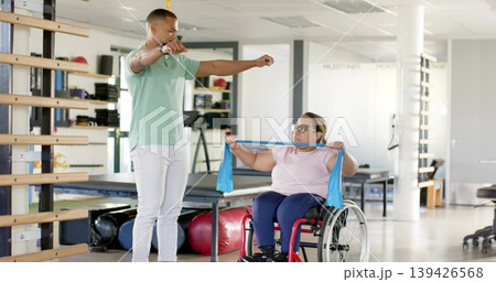 Guiding therapist in mint polo showing patient pulling blue band in rehab gym, red wheelchair 139426568