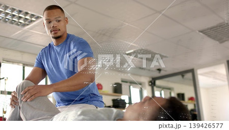 Supporting therapist wearing blue tee moving patient's leg on therapy table in clinic with med ball Supporting therapist wearing blue tee moving patient's leg on therapy table in clinic with med ball 139426577