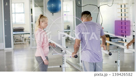 Practicing patient in purple tee and therapist in pink top walking between parallel bars at rehab Practicing patient in purple tee and therapist in pink top walking between parallel bars at rehab 139426582