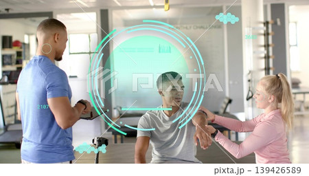 Seated client receiving forearm check in gym studio, trainers in athletic shirts using tablet HUD Seated client receiving forearm check in gym studio, trainers in athletic shirts using tablet HUD 139426589