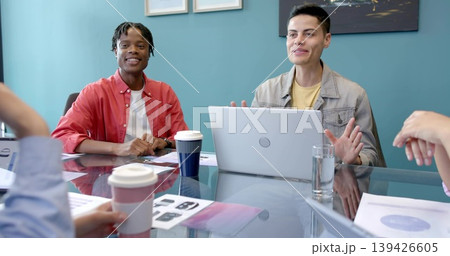 Discussing two colleagues in red button up and denim jacket at glass conference table, with laptop Discussing two colleagues in red button up and denim jacket at glass conference table, with laptop 139426605