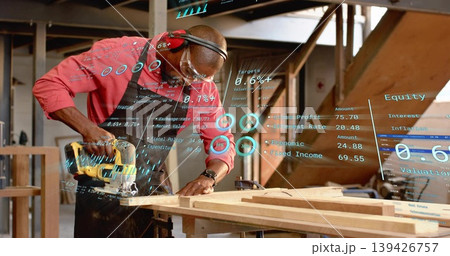 Carpenter wearing safety gear cutting wooden plank with circular saw in workshop with digital data 139426757