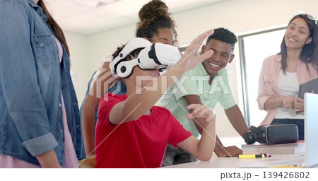 Reaching boy wearing white VR headset interacting at classroom table with instructor holding laptop Reaching boy wearing white VR headset interacting at classroom table with instructor holding laptop 139426802
