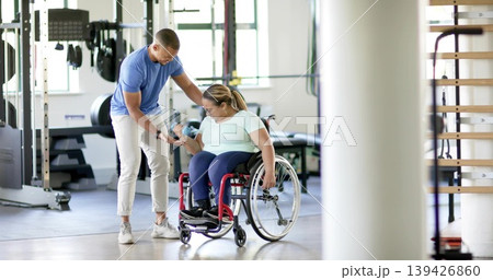 Guiding trainer steadying woman wearing active gear lifting blue dumbbell at gym, red wheelchair 139426860