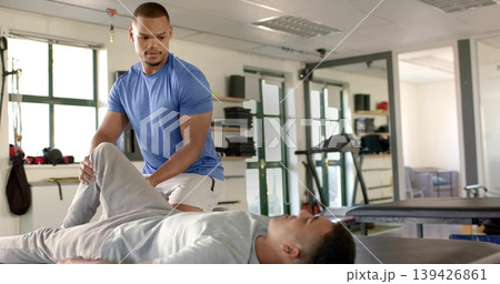 Kneeling trainer in blue T-shirt guiding bent leg on treatment table at gym with windows Kneeling trainer in blue T-shirt guiding bent leg on treatment table at gym with windows 139426861