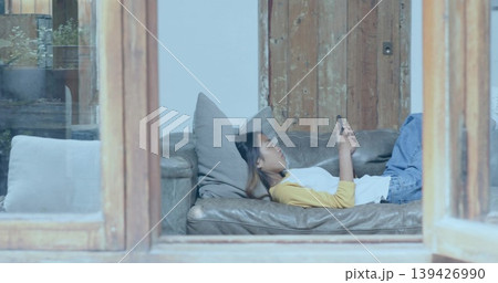 Reclining Asian woman in yellow top, jeans holding phone on sofa at home through wooden window Reclining Asian woman in yellow top, jeans holding phone on sofa at home through wooden window 139426990