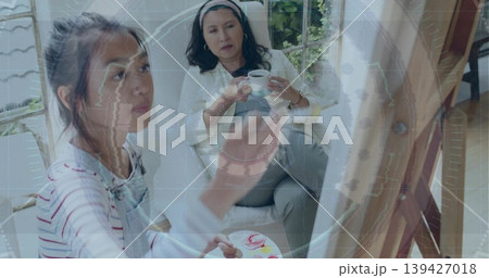 Painting artist wearing striped top and overalls at easel in sunroom, holding palette and brush Painting artist wearing striped top and overalls at easel in sunroom, holding palette and brush 139427018