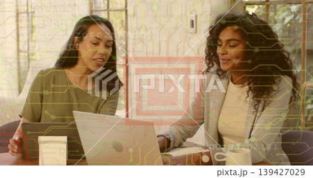 Talking two women in olive-tee and gray-blazer leaning over laptop at office holding tablet and mug Talking two women in olive-tee and gray-blazer leaning over laptop at office holding tablet and mug 139427029