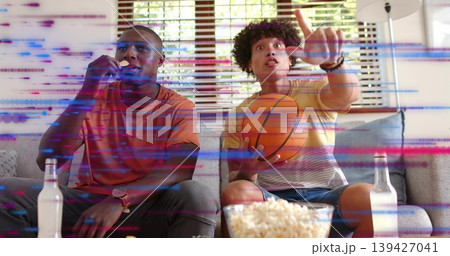 Watching men in orange, striped shirts on gray couch in living room, holding basketball and popcorn Watching men in orange, striped shirts on gray couch in living room, holding basketball and popcorn 139427041