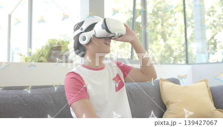Adjusting Asian man wearing white VR headset, red raglan tee in bright lounge, with mustard pillow 139427067
