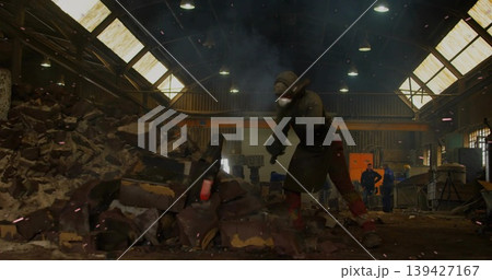 Striking worker wearing helmet mask apron gloves boots prying rubble at warehouse, handheld tool 139427167