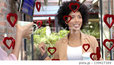 Talking woman tan blazer holding takeaway cup gesturing at mug with red heart overlays on patio 139427169