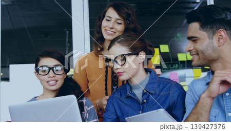 Collaborating team leaning over laptop and tablet at office in casual clothes, glass labeled CoLAB 139427376