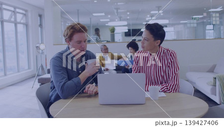 Talking pair wearing business-casual at round table in office lounge holding laptop and mugs 139427406