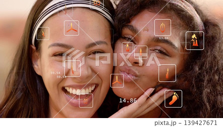 Laughing two adult women pressing cheeks together in studio, wearing headbands with HUD icons 139427671