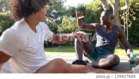 Sitting two men in activewear chatting and gesturing on wooden deck in backyard, with green bottle Sitting two men in activewear chatting and gesturing on wooden deck in backyard, with green bottle 139427689