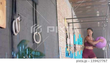 Holding purple medicine ball, maroon clad woman rotating in gym with rings and charts, copy space Holding purple medicine ball, maroon clad woman rotating in gym with rings and charts, copy space 139427767