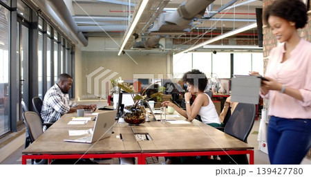 Working three coworkers sharing long table in open office, in casual attire, using laptops, tablet Working three coworkers sharing long table in open office, in casual attire, using laptops, tablet 139427780