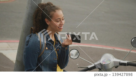 Leaning woman in denim jacket, yellow top holding phone at curb with scooter backpack, copy space Leaning woman in denim jacket, yellow top holding phone at curb with scooter backpack, copy space 139427898