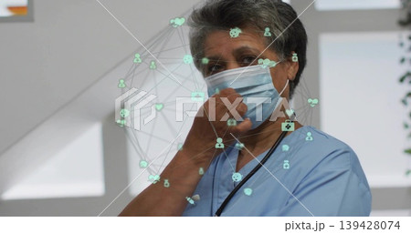 Adjusting mature clinician in blue scrubs holding mask in clinic, with stethoscope, digital network Adjusting mature clinician in blue scrubs holding mask in clinic, with stethoscope, digital network 139428074