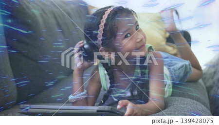 Lying girl in tank top and denim shorts on grey couch, holding tablet, pressing over-ear headphone Lying girl in tank top and denim shorts on grey couch, holding tablet, pressing over-ear headphone 139428075