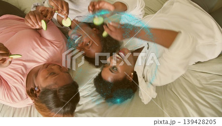 Lying three African American women in loungewear on bed, holding cucumber slices and blue glitter Lying three African American women in loungewear on bed, holding cucumber slices and blue glitter 139428205
