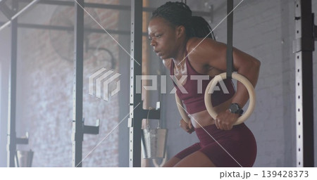 Holding woman wearing maroon sports bra performing ring dips on wooden rings in gym, copy space Holding woman wearing maroon sports bra performing ring dips on wooden rings in gym, copy space 139428373