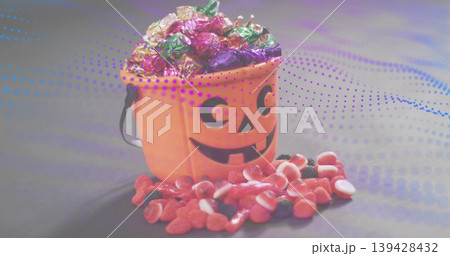 Displaying orange jack-o-lantern bucket holding foil-wrapped sweets on gray table, spilling gummies 139428432