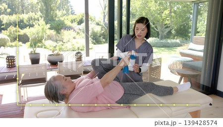 Holding blue band, pink-top senior lifting leg on patio therapy table, caregiver in scrubs guiding Holding blue band, pink-top senior lifting leg on patio therapy table, caregiver in scrubs guiding 139428574