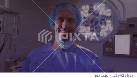 Standing surgeon in blue scrubs, bouffant cap and lowered mask under lights in operating room Standing surgeon in blue scrubs, bouffant cap and lowered mask under lights in operating room 139428618